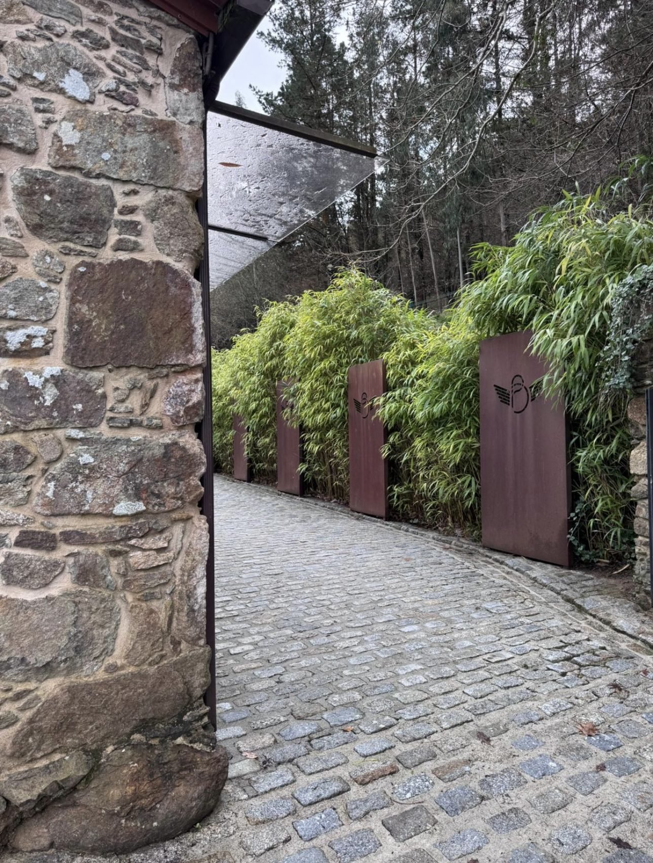 Camino empedrado de acceso entre muros de piedra y paneles de acero corten